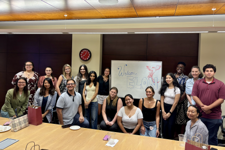 A group of students and faculty pose and smile in front of a whiteboard sign that reads “Welcome Elon!” in a conference room.